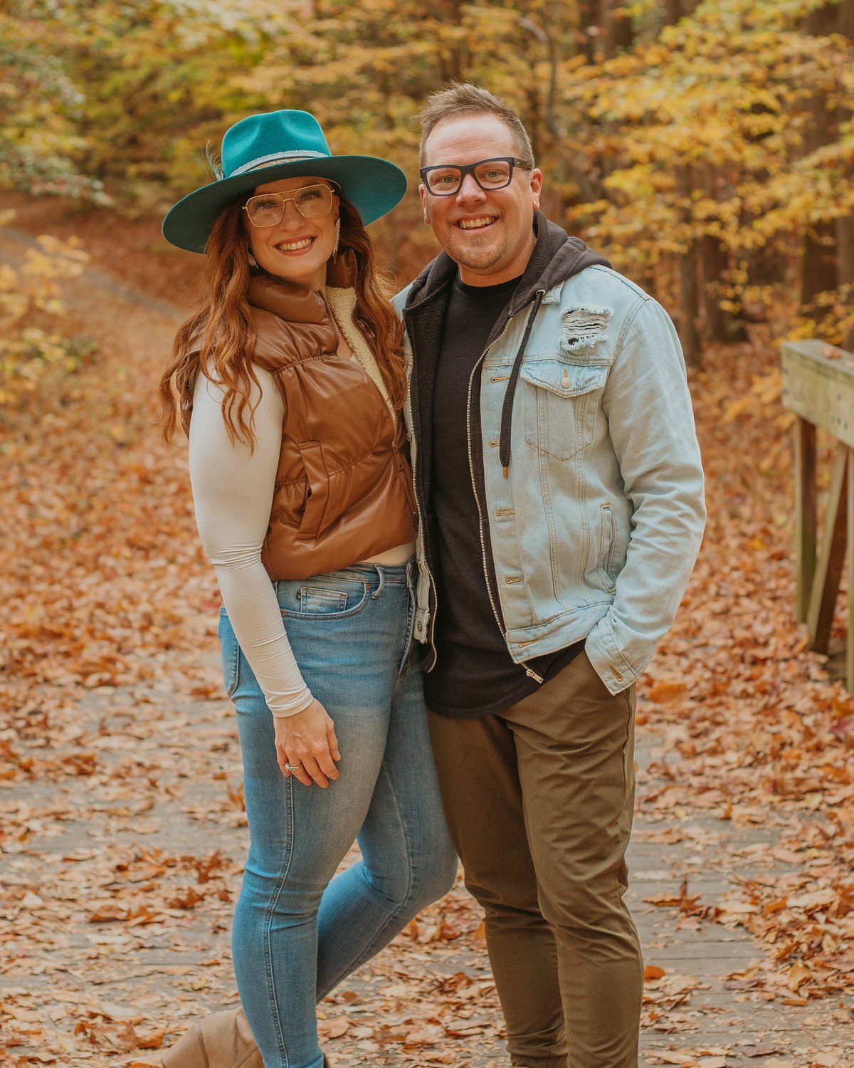 Shelly and Jason smiling on a wooden bridge in an autumn forest, Shelly in a teal hat
